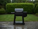 Black Traeger Westwood pellet grill on wet gravel, lid closed, hopper at right, clipped hedge behind