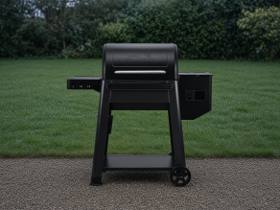 Black Traeger Westwood pellet grill on wet gravel, lid closed, hopper at right, clipped hedge behind