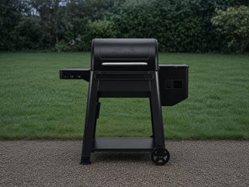 Black Traeger Westwood pellet grill on wet gravel, lid closed, hopper at right, clipped hedge behind