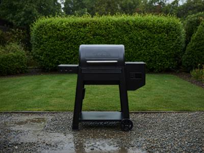 Black Traeger Westwood pellet grill on wet gravel, lid closed, hopper at right, clipped hedge behind