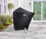 Covered Weber Performer grill on grey patio tiles beside a planted bed and a glass sliding door