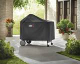 Covered Weber Performer on a paved patio beside a brick house, potted red flowers and green lawn