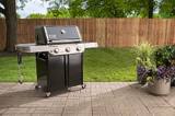 Weber Genesis E-315 on a paver patio beside a lawn, wooden fence and potted plants behind it