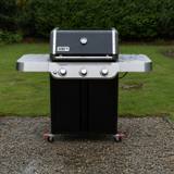 Weber Genesis E-315 front-on on wet gravel, lid closed, three knobs on a stainless panel, red caster locks visible