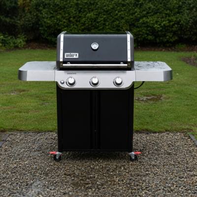 Weber Genesis E-315 front-on on wet gravel, lid closed, three knobs on a stainless panel, red caster locks visible