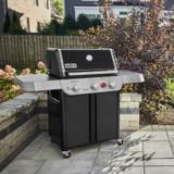 Genesis E-325 on a paved patio beside a low stone wall, shrubs behind, lid closed in afternoon light