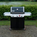 Black Weber Genesis E-325 gas grill on wet gravel beside a lawn, lid closed, hedge behind