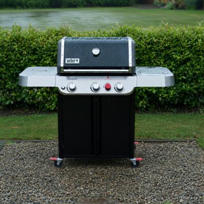 Black Weber Genesis E-325 gas grill on wet gravel beside a lawn, lid closed, hedge behind