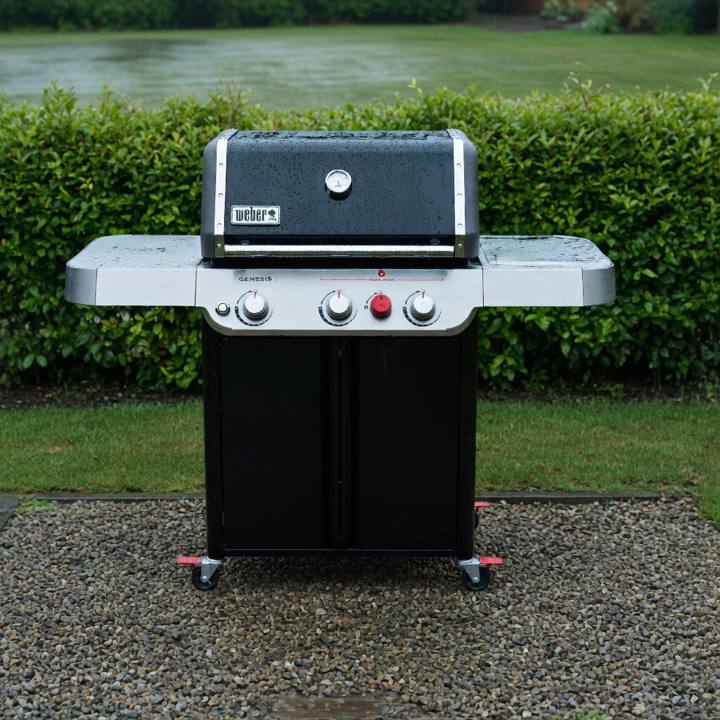 Black Weber Genesis E-325 gas grill on wet gravel beside a lawn, lid closed, hedge behind
