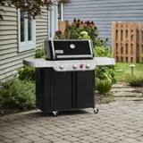 Weber Genesis E-330 on a paver patio beside a gray clapboard house, lid closed, cabinet cart on casters