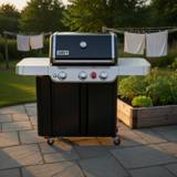 Black Weber Genesis gas grill on a stone patio at dusk, cabinet doors shut, stainless side tables, garden beds behind