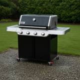 Weber Genesis E-415 four-burner propane grill, black lid beaded with rain, parked on wet gravel