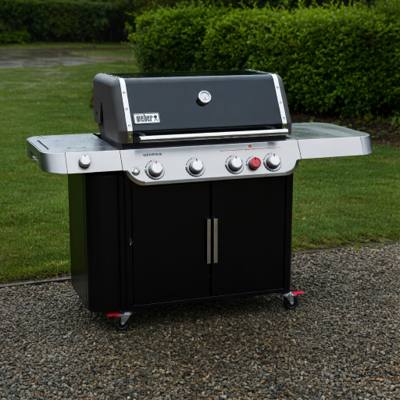 Black Weber Genesis E-435 gas grill on wet gravel, lid closed, red Sear Zone knob on the panel