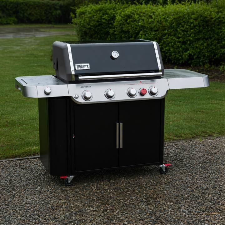 Black Weber Genesis E-435 gas grill on wet gravel, lid closed, red Sear Zone knob on the panel