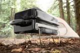 Weber Go-Anywhere charcoal grill steaming on its wire legs on a forest floor, a tent pitched behind it