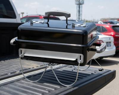Weber Go-Anywhere charcoal grill on a pickup tailgate in a stadium car park, wire legs folded down
