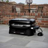 Weber Lumin electric grill on gray paving by a brick wall, lid closed, black body, bright metal handle, cord coiled beside it