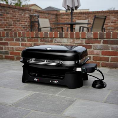 Weber Lumin electric grill on gray paving by a brick wall, lid closed, black body, bright metal handle, cord coiled beside it