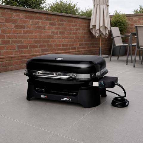 Weber Lumin electric grill on gray paving by a brick wall, lid closed, black body, bright metal handle, cord coiled beside it