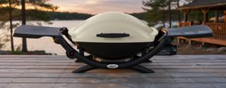 Weber Q 2000 portable gas grill on a lakeside deck, cream lid closed and both side tables folded out, viewed from the front
