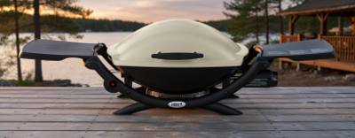 Weber Q 2000 portable gas grill on a lakeside deck, cream lid closed and both side tables folded out, viewed from the front
