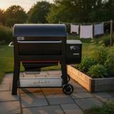 Black Weber Searwood pellet grill on flagstones by a raised vegetable bed, hopper and lit controller on the right