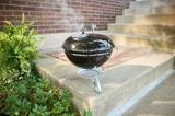 Weber Smokey Joe 14 standing on concrete steps beside a brick wall, lid closed, smoke drifting from the damper