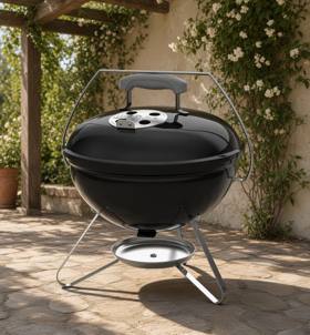 Black Weber Smokey Joe 14 kettle on a patio, lid on, chrome bail handle raised, silver ash catcher below the tripod legs