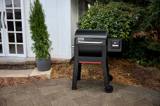 Weber Smoque pellet smoker on a paved patio beside a house door, conifer in a planter and hedge behind