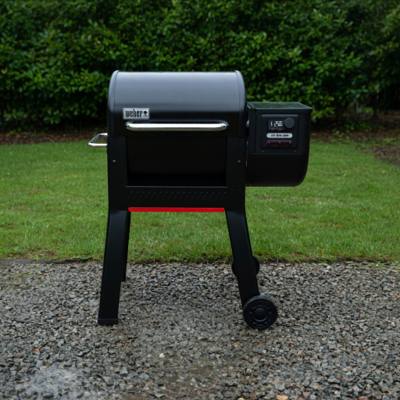 Weber Smoque pellet smoker on gravel beside a lawn, lid closed, side hopper and LCD controller to the right