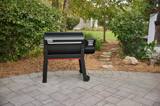 Weber Smoque XL standing on a paved patio with shrubs behind it, lid closed, seen from the front left