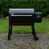 Black Weber Smoque XL pellet smoker on gravel beside a lawn, lid closed, lit controller on the right hopper, red stripe below