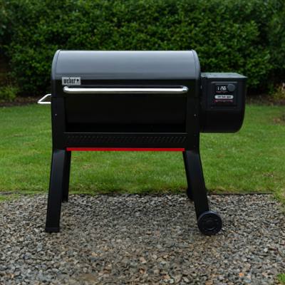 Black Weber Smoque XL pellet smoker on gravel beside a lawn, lid closed, lit controller on the right hopper, red stripe below