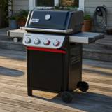 Weber Spirit E-435 on a timber deck at dusk, lid closed, four panel knobs and the side burner knob at left