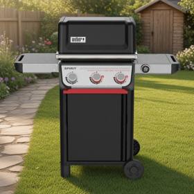 Black Weber Spirit gas grill, lid closed, on a mown lawn beside a paved path, wooden fence and shed behind