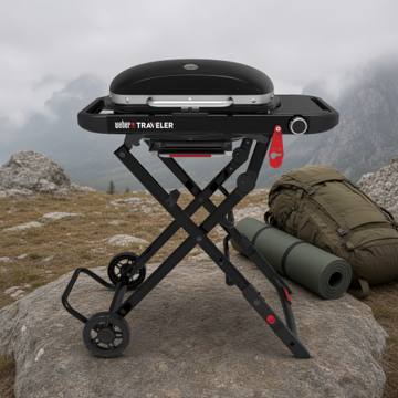 Black Weber Traveler Compact open on its folding X-frame on a rock, lid closed, rucksack and rolled mat behind