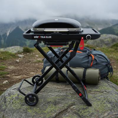 Black Weber Traveler Compact open on its folding X-frame on a rock, lid closed, rucksack and rolled mat behind
