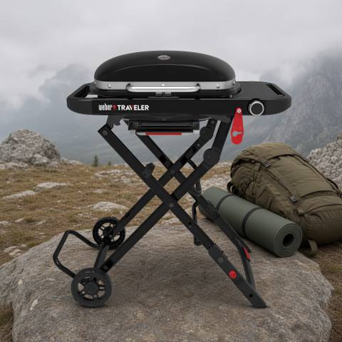 Black Weber Traveler Compact open on its folding X-frame on a rock, lid closed, rucksack and rolled mat behind