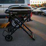 Weber Traveler portable gas grill standing on its folding frame behind a van, lid closed and side table out