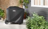 Covered Weber Traveler standing on a concrete patio beside a brick wall, potted plants and a green shrub